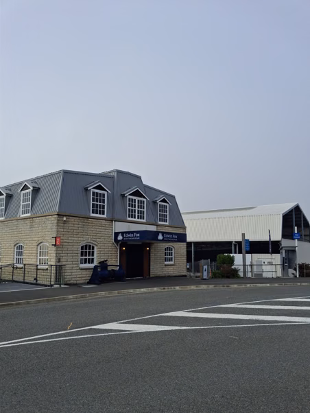 Colour photograph of the Edwin Fox Museum building on the left, with the shed housing the ship’s remains partly visible behind it on the right.